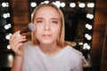 Serious young blonde woman look on camera. She stand in beauty room. Applying blush on cheeks using brush. Mirror with Royalty Free Stock Photo