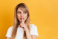 Serious young blonde woman posing over yellow wall background dressed in white casual t-shirt showing silence gesture