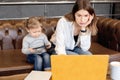 Serious young businesswoman in maternity leave sitting on sofa with small kid, using laptop, making important reports Royalty Free Stock Photo