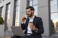 Serious young european man manager with beard in suit watch video on laptop with cup of coffee takeaway, eats croissant