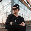 Serious young hipster man in black fashionable clothes in a knitted hat in trendy sunglasses is posing standing in a city Royalty Free Stock Photo