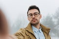 Serious young male is making self portrait on camera outdoors. Handsome traveler man wear coat, blue shirt and eyeglasses making Royalty Free Stock Photo