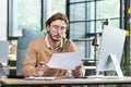 A serious young male programmer sits in the office at a table, works at a computer and documents. He holds a project, a