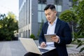 Serious young man, Asian businessman working outside an office building using a laptop and making notes in a notebook Royalty Free Stock Photo