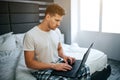 Serious young man in bed this morning. He work at home. Guy type on laptop keyboard and look at screen. Daylight.