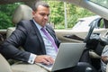 Serious young man in a business suit sits in the driver`s seat in the car with laptop. Busy businessman with short hair