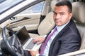 Serious young man in a business suit sits in the driver`s seat in the car with laptop. Busy businessman with short hair
