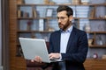 Serious young man businessman lawyer in suit and glasses standing in office and holding laptop, working, typing on Royalty Free Stock Photo