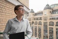 Serious young man in glasses standing on an open-air balcony with laptop, having break and looking thoughtfully. Flexible hours Royalty Free Stock Photo