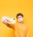 Serious young man in a mask against coronavirus in a yellow sweater makes a selfie on a smartphone on a yellow background,