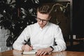 Serious young man wearing white shirt and eye glasses sitting at the table holding pen takes notes to his agenda on a dark Royalty Free Stock Photo