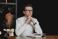 Serious young man wearing white shirt and eye glasses sitting at the table holding pen takes notes to his agenda on a dark Royalty Free Stock Photo