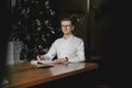 Serious young man wearing white shirt and eye glasses sitting at the table holding pen takes notes to his agenda on a dark Royalty Free Stock Photo