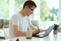 Serious young man working online with laptop at home, holding paper sitting at office desk Royalty Free Stock Photo