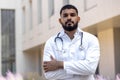 Serious young muslim male doctor standing outside clinic outside wearing a robe and holding a stethoscope and crossing Royalty Free Stock Photo