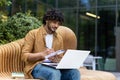 Serious young Muslim man sitting outside on a bench wearing headphones and using a laptop, writing in a notebook Royalty Free Stock Photo