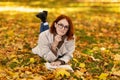 Serious young smart european red-haired lady in glasses and raincoat lies on yellow leaves, reads book