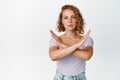 Serious young woman shows cross stop gesture, looking unamused, saying no, forbid something, standing over white