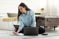 Serious young woman sitting on floor, working on laptop, taking notes in notebook in bedroom Royalty Free Stock Photo