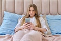 Serious young woman with smartphone resting lying at home in bed, reading surprised focused
