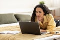 Serious young woman using laptop, working or studying online, having business meeting or webinar, lying on bed at home Royalty Free Stock Photo