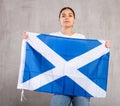 Serious young woman waving state flag of Scotland against gray wall background