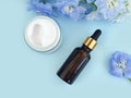 Serum in unbranded brown glass bottle with pipette, moisturizer cream in open glass jar and blue delphinium flower on blue