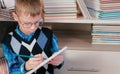 Seven-year-old boy with glasses thinking and draws something in a sketchbook sitting among the books. Royalty Free Stock Photo