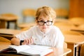 Seven years old child with glasses writing his homework at school. Boy studing at table on class background