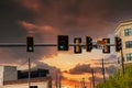 Several black and yellow traffic signals on a pole surrounded by buildings, lush green trees and cars with powerful red clouds