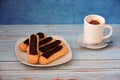 Several chocolate-coated cream eclairs on a plate and a cup of hot cappuccino on a wooden table