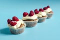 Several cupcakes with white cream and raspberries lined up on a blue background sharpness in the foreground.