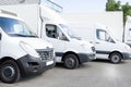 Several white row commercial delivery vans and service van, trucks and car in front of factory warehouse