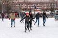 Several young and old people skating at a public ice skating rink outdoors in the city.