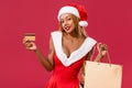 African american girl in santa hat and christmas dress holding credit card and shopping bag isolated on red Royalty Free Stock Photo