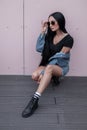 Sexy american young hipster woman in trendy leather boots in a denim jacket in sunglasses in a skirt sits outdoors Royalty Free Stock Photo