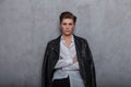 Sexy attractive young man model in a fashionable leather black jacket in an elegant white classic shirt poses in a studio near a