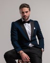 sexy bearded groom in elegant tuxedo looking forward and sitting with hand in pocket