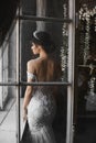 and beautiful brunette model girl in lace white dress with naked shoulders with diadem on her head stands near the window and Royalty Free Stock Photo