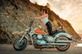 Sexy beautiful woman in high heels and leather pants posing on motorcycle. Mountain and sunset sky at the background