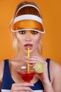 Sexy woman in swimsuit and trendy plastic visor holding glass of fresh drink on yellow background