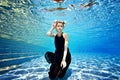 Sexy blonde girl sits underwater at the bottom of an outdoor pool in a black dress with white horns on her head on a