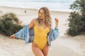 blonde girl in a yellow swimsuit and glasses on her face, holding a denim jacket, having fun on the beach with Royalty Free Stock Photo