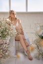 Sexy blonde woman in a beautiful white dress is sitting near the window in front of a bouquet of wild flowers. Romantic girl with