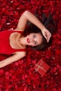 A girl in a red dress, lies in the rose petals near a gift box and holds her hand near the head. Royalty Free Stock Photo