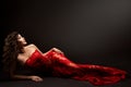 Sexy Brunette Woman In Red Lace Dress lying down over Black. Fashion Model in Long Evening Gown with Curly Hair looking up Happy Royalty Free Stock Photo