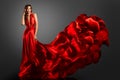 Sexy Fashion Model in Red Silk Dress. Glamour Woman in Long Luxury Gown flying on Wind with Wavy Hairstyle over Dark Gray Royalty Free Stock Photo