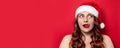 Sexy girl with red santa hat with pompom licks lips on red wall background