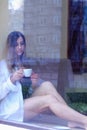 Sexy girl sitting near the window and drinking coffee. single woman in white men`s shirt, sad and thinks