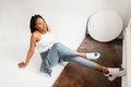 Sexy modern African woman with dreadlocks in white youth top in blue stylish jeans in trendy sneakers sits on floor indoor